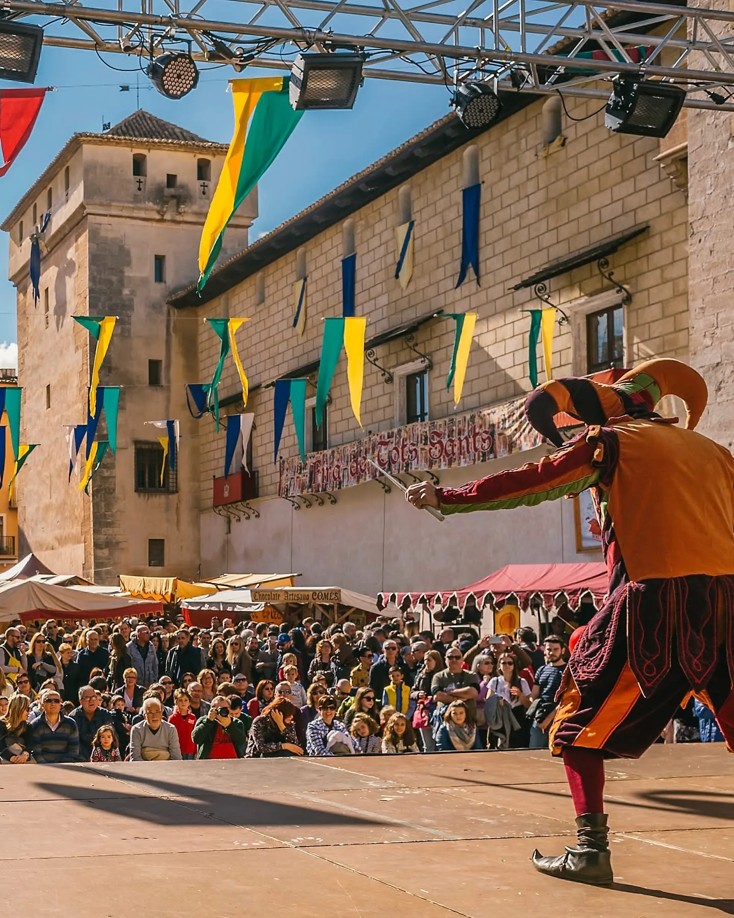 Live Performances at the Medieval Stage in Cocentaina A costumed performer entertains a crowd on an outdoor stage during the Fira de Tots Sants, with medieval-style banners hanging above.