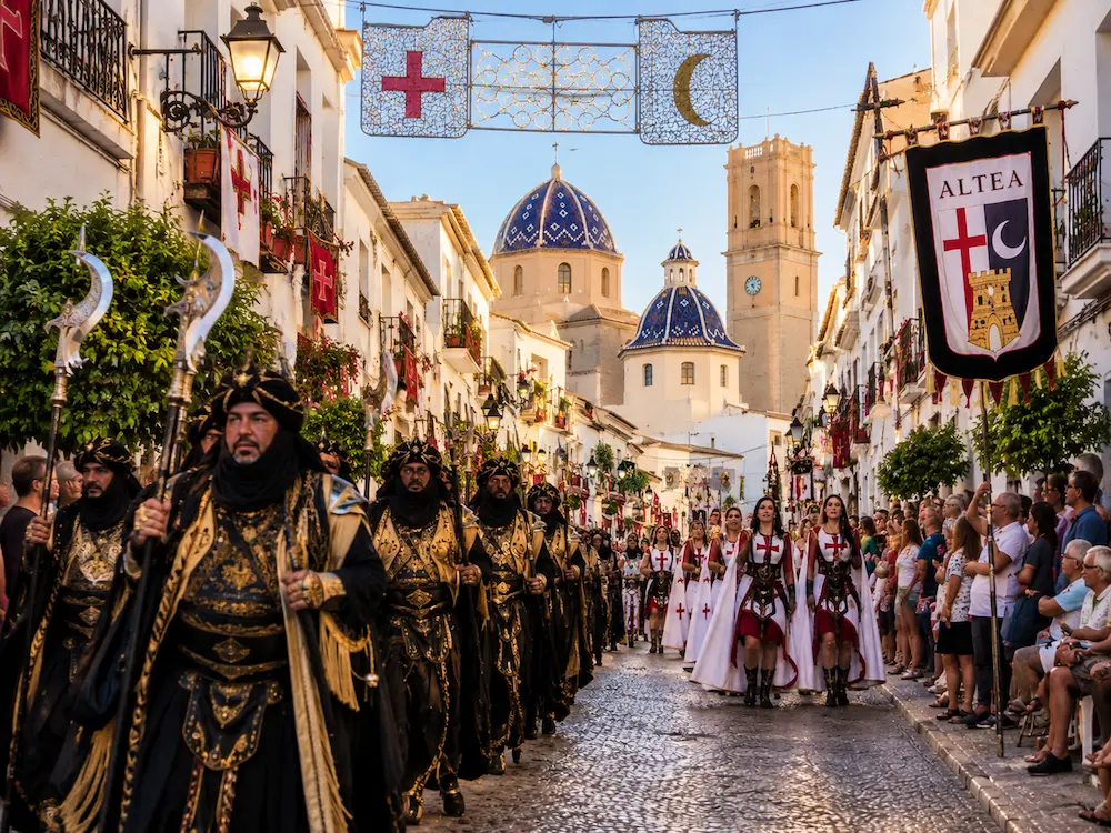 Moros y Cristianos Festival in Altea Moros y Cristianos parade through the historic streets of Altea, with Moorish and Christian-costumed participants, festive banners and decorations, and Altea’s iconic blue-tiled church domes in the background.
